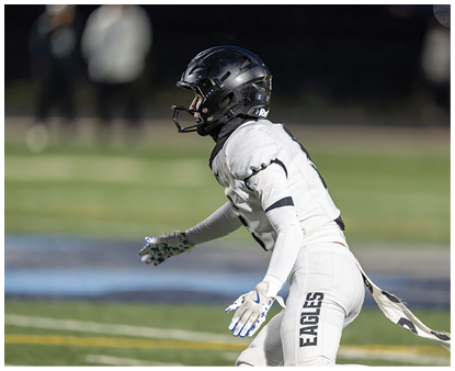 A person wearing a white football uniform with “EAGLES" on the pants and a black helmet is in an athletic stance on a football field. Description generated by AI