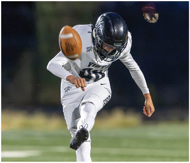 A person wearing a white sports uniform and black helmet is kicking a football on a field. Description generated by AI