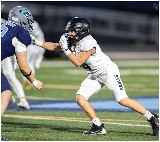 Two people in football uniforms and helmets are engaged in a play on a field, with one person in white lunging forward and extending an arm toward the other person in blue. Description generated by AI