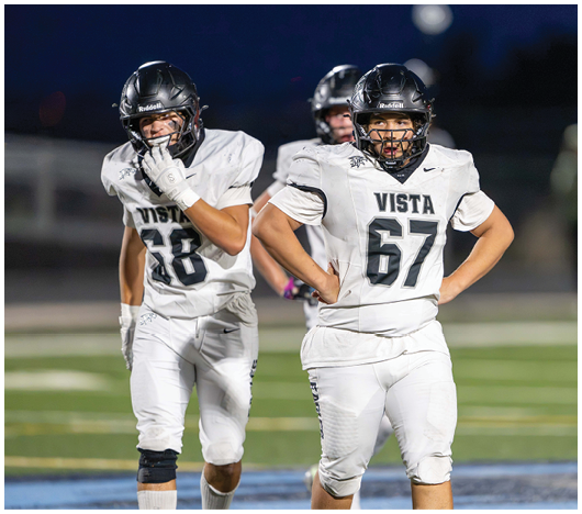 Two people in football uniforms with helmets stand on a field at night, one with hands on hips and the other with a hand near their mouth. Description generated by AI