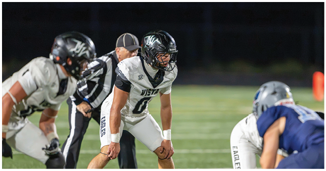 People in football uniforms are positioned on a field preparing for a play during a game. Description generated by AI