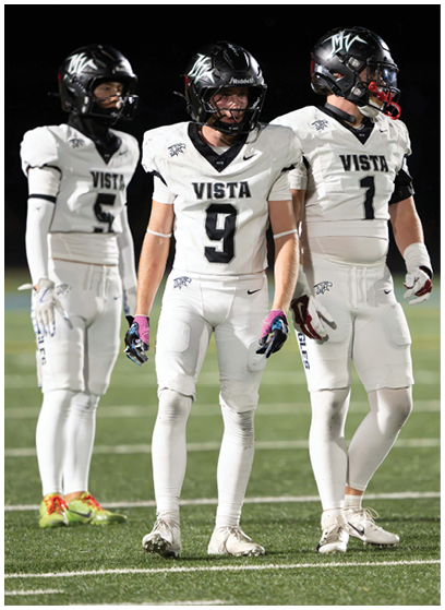 Three people in white football uniforms with black helmets stand on a field under bright lights during a game. Description generated by AI