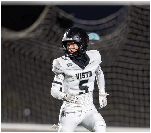 A person wearing a black helmet and white football uniform with the word Vista and the number 5 is running on a field with a goal net in the background. Description generated by AI