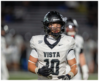 A person wearing a white football uniform with the word Vista and the number 10 on the front is standing on a field with a black helmet and gloves. Description generated by AI