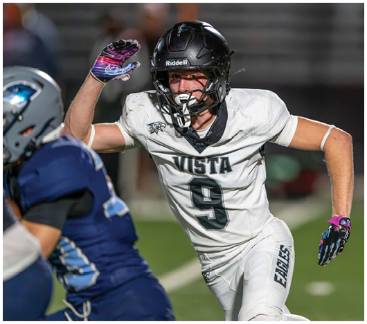 A person in a white football uniform with the number 9 and the word Vista is running on a field while raising one arm, facing another person in a blue football uniform. Description generated by AI