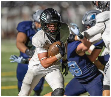 A person in a white football uniform is running with the ball while being pursued and tackled by players in blue uniforms during a game. Description generated by AI