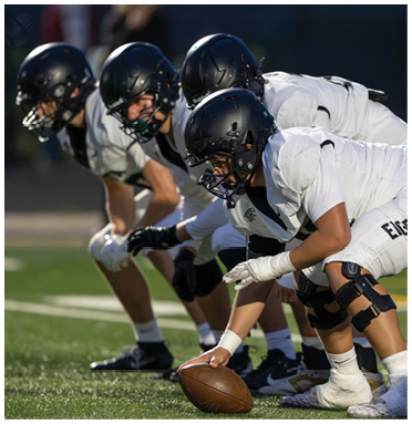 Four people in football helmets and white uniforms are lined up on a field with one person holding a football ready to snap it. Description generated by AI