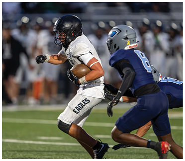 A person in a white football uniform with “EAGLES" on the pants is running with the ball while being pursued by a person in a blue football uniform on a field. Description generated by AI