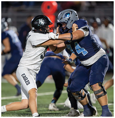 Two people in football uniforms and helmets are engaged in a physical block during a game on a field. Description generated by AI