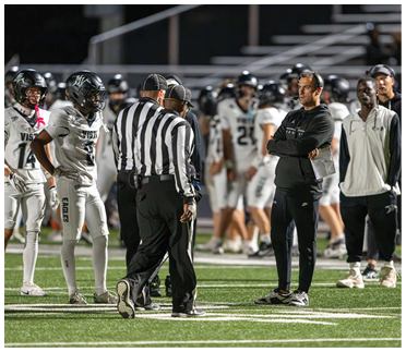 People in football uniforms and referees are gathered on a field with coaches standing nearby during a game. Description generated by AI