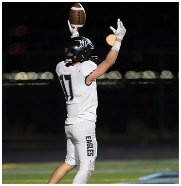 A person wearing a white football uniform and black helmet is raising a football above their head on a field at night. Description generated by AI