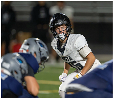 A person in a white football uniform and black helmet is crouched in a ready position facing two people in blue uniforms and silver helmets on a football field. Description generated by AI