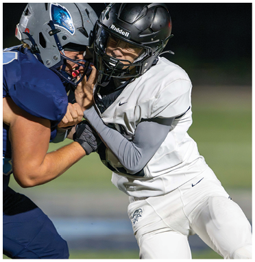 Two people wearing football helmets and uniforms are engaged in a physical play on a football field at night. Description generated by AI