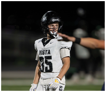 A person wearing a white football uniform with the number 35 and a black helmet is standing on a field looking to the side while another person’s arm is extended in front of them. Description generated by AI