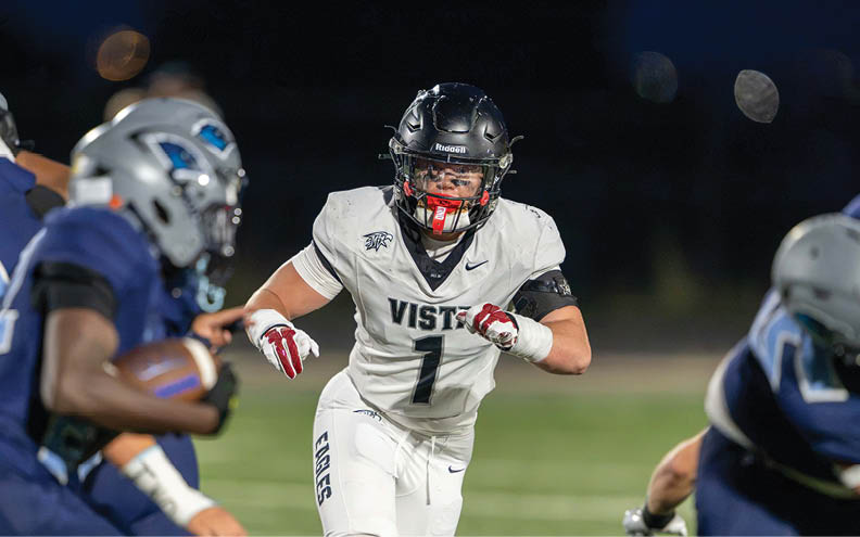 A person in a white football uniform with the word Vista is running on a field toward a person in a blue football uniform holding a football. Description generated by AI