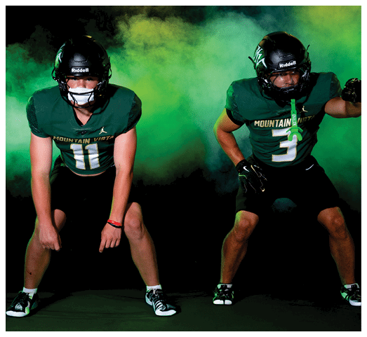 Two people in green football uniforms and black helmets are posed in a ready stance against a dark background with green smoke. Description generated by AI