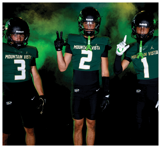 Two people wearing Mountain Vista football uniforms with helmets and gloves pose against a dark background with green smoke, one holding up three fingers and the other holding up two fingers. Description generated by AI