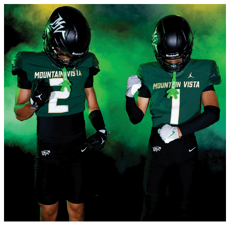 Two people wearing black and green football uniforms with helmets stand against a smoky green and yellow background, with one person looking down at their chest. Description generated by AI