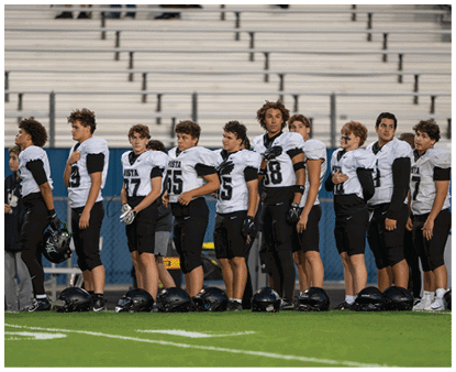 A group of people wearing white and black football uniforms stand in a line on a field with helmets placed on the ground in front of them and empty bleachers in the background. Description generated by AI