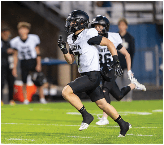 A person wearing a black helmet and white sports jersey with the word Vista is running on a football field with other players in the background. Description generated by AI