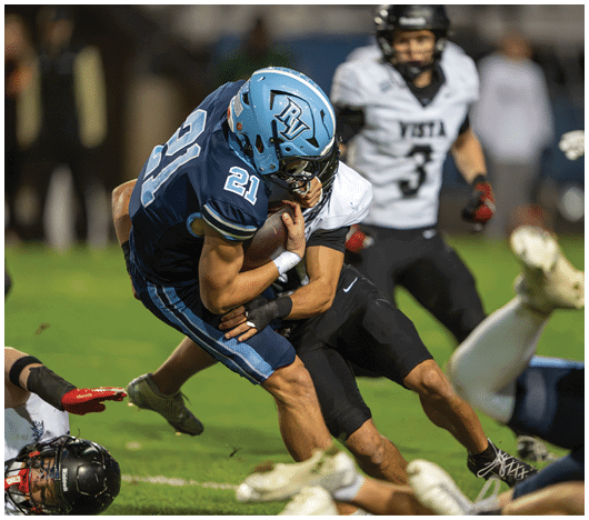 Two people in football uniforms are engaged in a tackle during a game, with one player wearing a blue helmet and jersey numbered 21 holding the ball. Description generated by AI