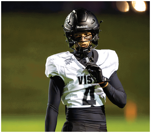 A person wearing a black helmet and white football jersey with the number 4 is standing on a field holding a microphone near their mouth. Description generated by AI