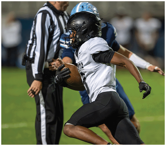 A person in a white and black football uniform is running with a football while another person in a blue uniform and a referee stand nearby on a grassy field. Description generated by AI