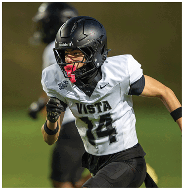 A person wearing a black helmet and white football jersey with the number 14 is running on a field during a game. Description generated by AI