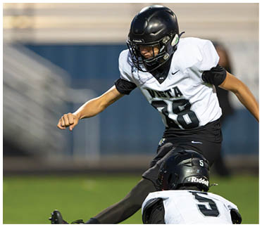 Two people in football uniforms are on a field with one person holding the ball on the ground while the other is kicking it. Description generated by AI