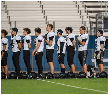 A line of people in football uniforms stand side by side on a field with their helmets on the ground in front of them, some holding their hands over their chests. Description generated by AI