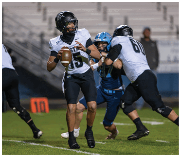 A person in a black and white football uniform is holding a football and preparing to throw while another person in a similar uniform blocks an opponent in a blue uniform on a football field. Description generated by AI