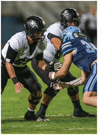 Two people in black and white football uniforms are lined up against a person in a blue football uniform on a grassy field during a game. Description generated by AI