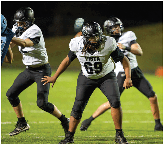 Three people in football uniforms and helmets are positioned on a grassy field, appearing ready to engage in a play during a game. Description generated by AI