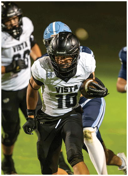 A person wearing a black helmet and white sports jersey with the number 10 is running with a football while being pursued by two people in blue sports uniforms on a grassy field. Description generated by AI