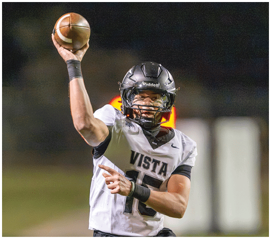 A person wearing a football helmet and a white jersey with the word Vista is preparing to throw a football during a nighttime game. Description generated by AI