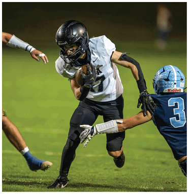 A person in black and white football gear is running with the ball while another person in blue football gear attempts to grab their arm on a grassy field. Description generated by AI