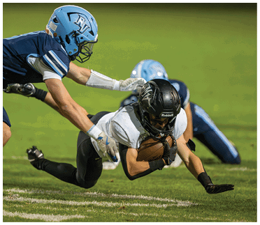 Two people in football gear are in mid-air on a grassy field with one person holding a football while the other reaches out to tackle. Description generated by AI