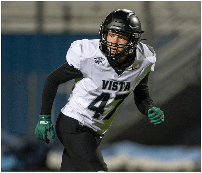 A person wearing a football helmet, white jersey with the word Vista and number 47, black pants, and green gloves is running on a field. Description generated by AI
