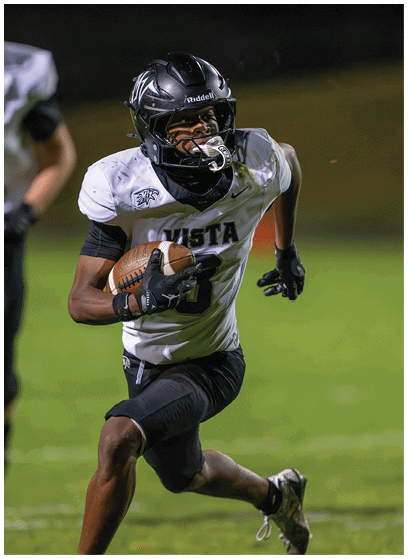 A person wearing a black helmet and white football jersey with “VISTA" on it is running on a grassy field while holding a football. Description generated by AI