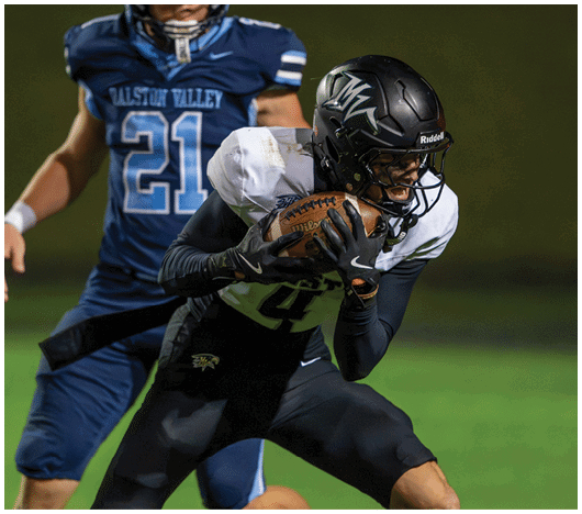 A person in black and white football gear is holding a football tightly while being closely pursued by another person in a blue uniform on a grassy field. Description generated by AI