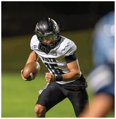 A person wearing a black helmet and white football jersey with the number 15 is running on a grassy field while holding a football. Description generated by AI