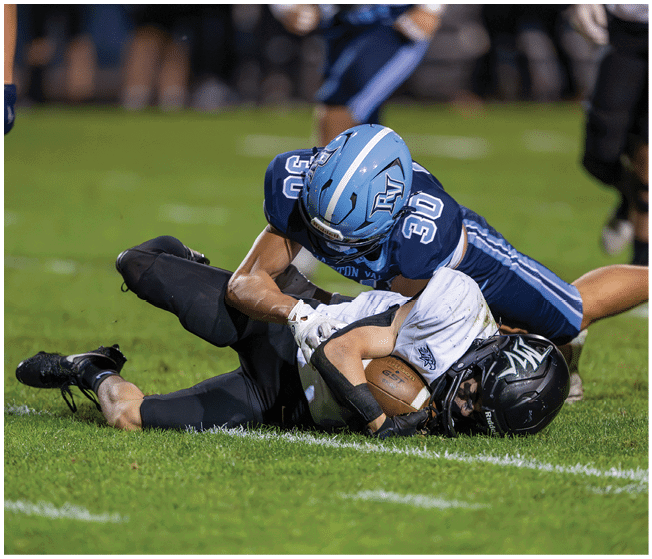 A person in a dark blue football uniform tackles another person in a white and black football uniform who is holding a football on a grassy field. Description generated by AI