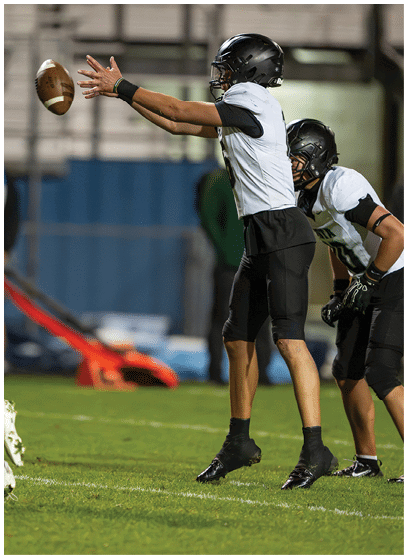 Two people in football uniforms are on a field with one person reaching out to catch a football while the other runs nearby. Description generated by AI