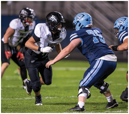 Two people in football uniforms and helmets are actively engaged on a grassy field with one person running toward the other who is in a defensive stance. Description generated by AI
