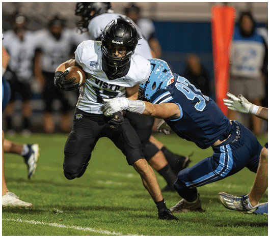 A person in a white and black football uniform is running with the ball while being tackled by a person in a blue football uniform on a grassy field during a game. Description generated by AI