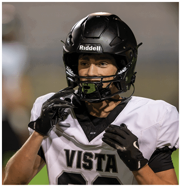 A person wearing a black football helmet and white jersey with the word Vista is adjusting their gloves on a sports field. Description generated by AI