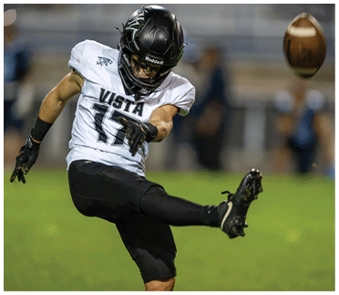 A person wearing a football uniform and helmet is kicking a football on a grassy field during a game. Description generated by AI