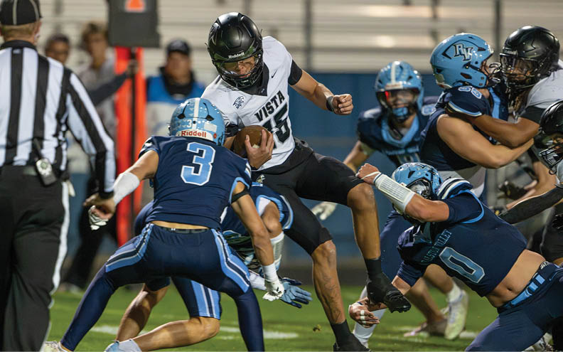 A group of people in football uniforms are actively engaged in a play on a field, with one person in a white jersey holding the ball while others in blue jerseys attempt to tackle or block. Description generated by AI