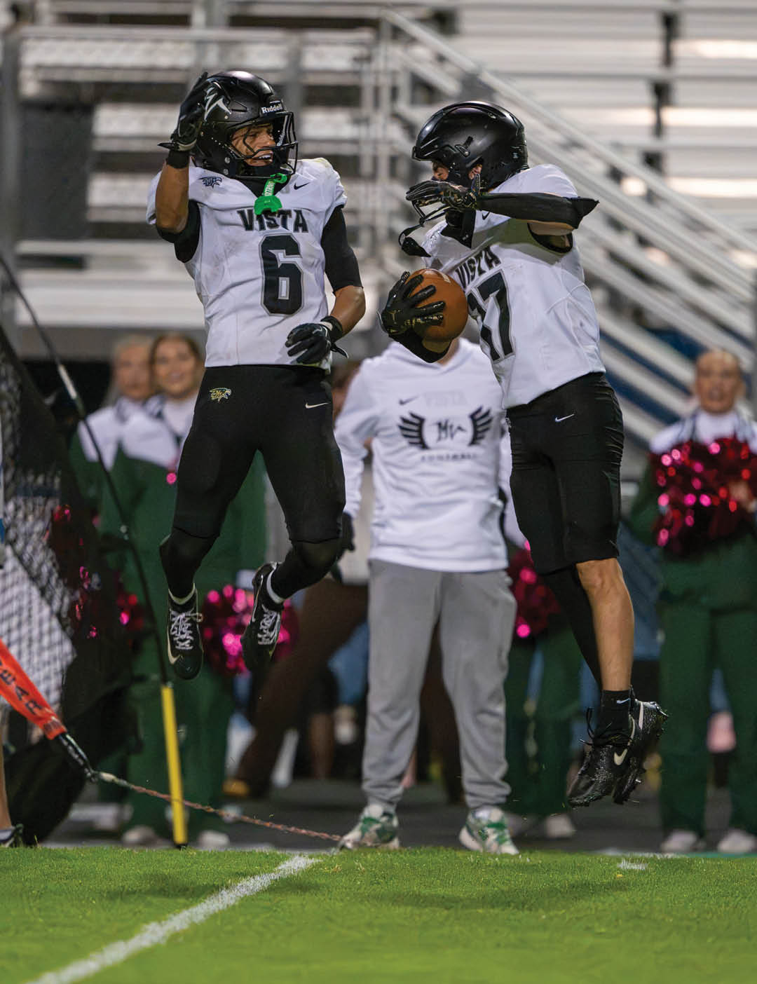 Two people in football uniforms celebrate on the field with one jumping in the air while others and a coach watch near the sideline. Description generated by AI