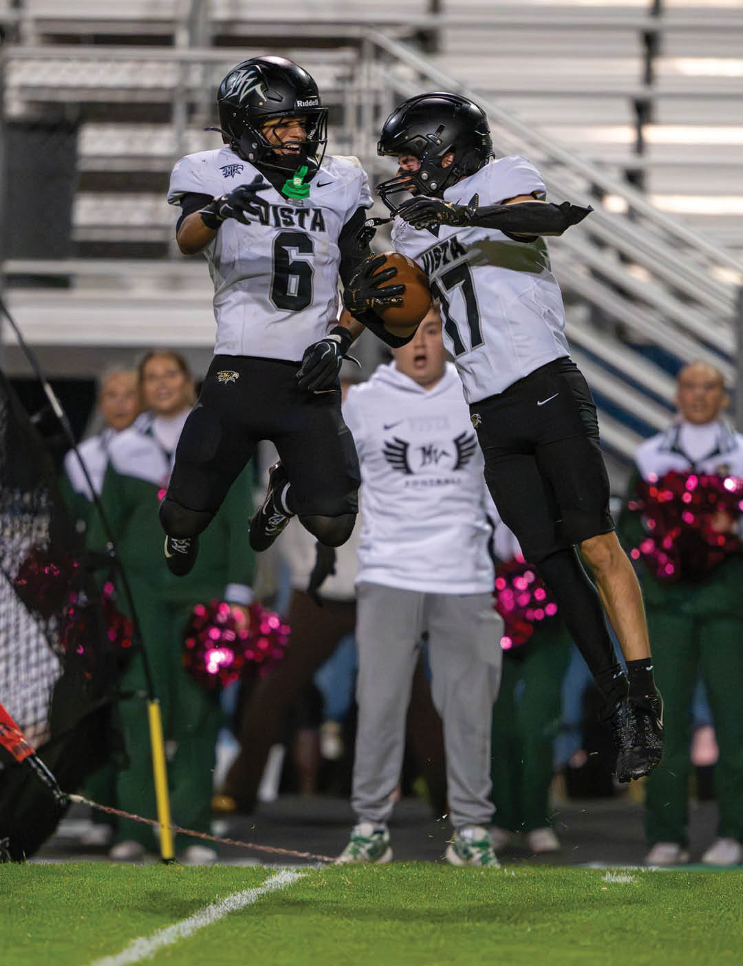 Two football players in black helmets and white jerseys jump in the air celebrating on a field with cheerleaders and spectators in the background. Description generated by AI